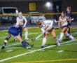 Women playing competitive field hockey at night in action-packed match on artificial turf.
