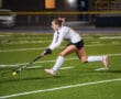 Field hockey player in action, mid-strike on a green turf field under stadium lights, focused and determined.