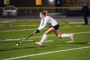 Field hockey player in action, mid-strike on a green turf field under stadium lights, focused and determined.