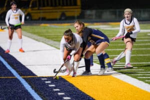 High school field hockey match at night, players competing for the ball on the sidelines.