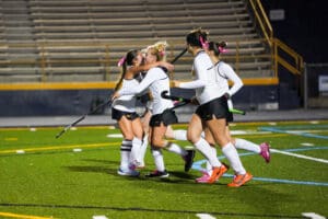 Field hockey players celebrating a goal on the field with joyful team embrace, wearing matching uniforms and gear.