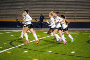 Field hockey players in action on a green field, wearing white and black uniforms with colorful shoes.