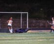 Field hockey players in action; goalie defending a shot near the goal, under stadium lights at night.
