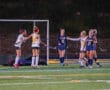 Girls' field hockey match with players celebrating a goal on a brightly lit turf field.