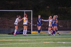Girls' field hockey match with players celebrating a goal on a brightly lit turf field.
