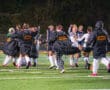 Field hockey team celebrates victory on the field at night, wearing jerseys and black jackets, showing team spirit.