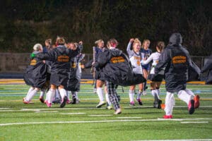 Field hockey team celebrates victory on the field at night, wearing jerseys and black jackets, showing team spirit.