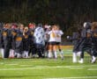Girls field hockey team celebrates victory at night on illuminated field, players hugging and wearing team jackets.