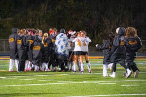 Girls field hockey team celebrates victory at night on illuminated field, players hugging and wearing team jackets.