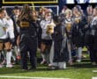 Field hockey team celebrates on the field, wearing uniforms and jackets, under the stadium lights.