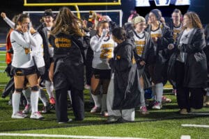 Field hockey team celebrates on the field, wearing uniforms and jackets, under the stadium lights.