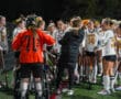 High school field hockey team in orange and white uniforms listens to coach during a night game.