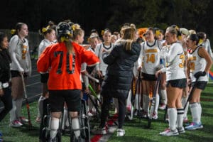 High school field hockey team in orange and white uniforms listens to coach during a night game.