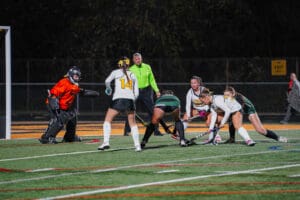 Girls' field hockey match at night with players competing for the ball near the goal.