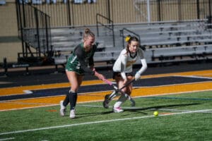Two female field hockey players compete for the ball on a vibrant sports field during a match.