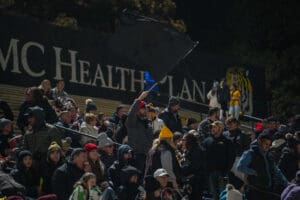 Crowd in stadium stands at night, person waving flag, others wearing winter clothing, large sign in background.