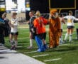 Field hockey team with mascot on sidelines during night game.