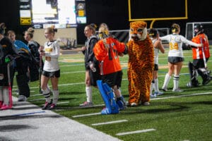 Field hockey team with mascot on sidelines during night game.