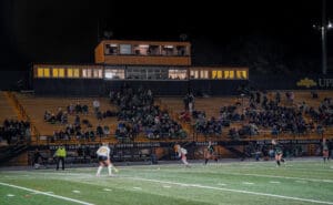 Field hockey match under lights, with spectators on bleachers and players in action, at North Allegheny stadium.