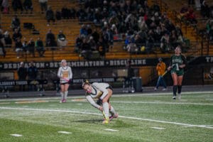 Field hockey player in action during a night game, with spectators watching in the stands.