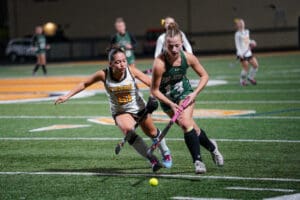Two field hockey players compete intensely for the ball on a green turf field.