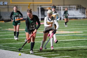 High school field hockey game, players in action on turf, focus on two athletes competing for the ball.
