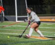 Field hockey player poised to hit the ball towards the goal on a brightly lit turf field.