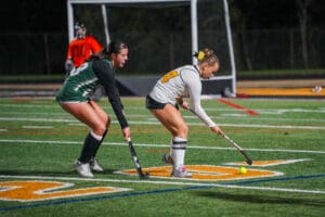 Two women playing field hockey at night, one in white and one in green, competing intensely on a lit field.
