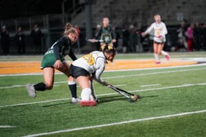 Two field hockey players competing under stadium lights on a turf field.