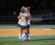 Field hockey players in white jerseys embrace on the field during a night game.