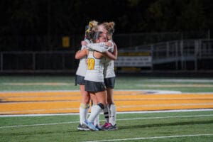 Field hockey players in white jerseys embrace on the field during a night game.