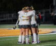 Field hockey team huddles on the field, wearing uniforms, expressing unity and teamwork before the game.