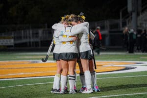 Field hockey team huddles on the field, wearing uniforms, expressing unity and teamwork before the game.