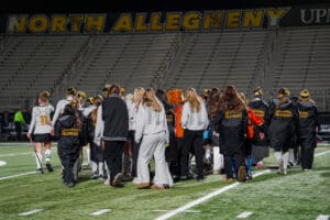 Field hockey team gathers on North Allegheny stadium field at night, wearing team jackets, facing bleachers.