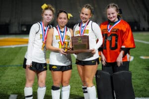 Girls' field hockey team with medals, holding a runner-up plaque on a sports field.