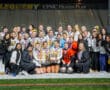 Group of athletes wearing medals, celebrating with a trophy on a sports field at night, smiling for the camera.