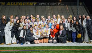 Group of athletes wearing medals, celebrating with a trophy on a sports field at night, smiling for the camera.