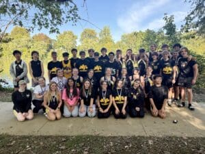 Group of young rowers posing by a sunny lakeside in team uniforms, surrounded by lush greenery.