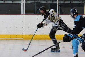 Roller hockey player in black and white uniform taking a shot, with a defender nearby on the rink.