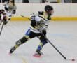 Roller hockey player in action on indoor rink, focusing on puck control with teammates in the background.