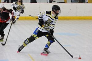 Roller hockey player in action on indoor rink, focusing on puck control with teammates in the background.