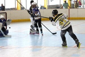 Player in roller hockey game shooting puck towards goal as goalie prepares to block, action-packed rink scene.