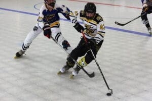 Youth roller hockey players compete on rink during a game, wearing colorful jerseys and helmets, with sticks and puck.