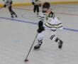 Youth roller hockey player in action on rink, wearing white jersey and helmet, with teammates in the background.