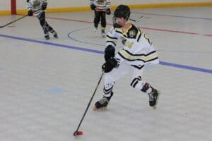 Youth roller hockey player in action on rink, wearing white jersey and helmet, with teammates in the background.