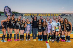 North Allegheny field hockey team poses on the field with their coach, holding posters of teammates in black uniforms.