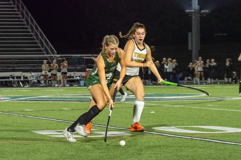 Two female field hockey players competing for the ball during a night game on a stadium field.