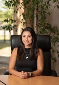 Smiling woman at office desk, sitting confidently, black top, indoor plant background.