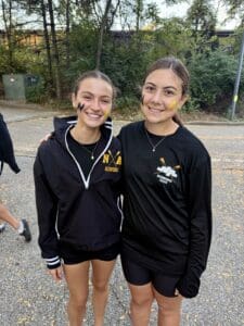 Two smiling women in rowing team gear with face paint, outdoors with trees in the background.