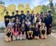 Smiling rowing team poses with medals by the river, surrounded by greenery and blue sky.